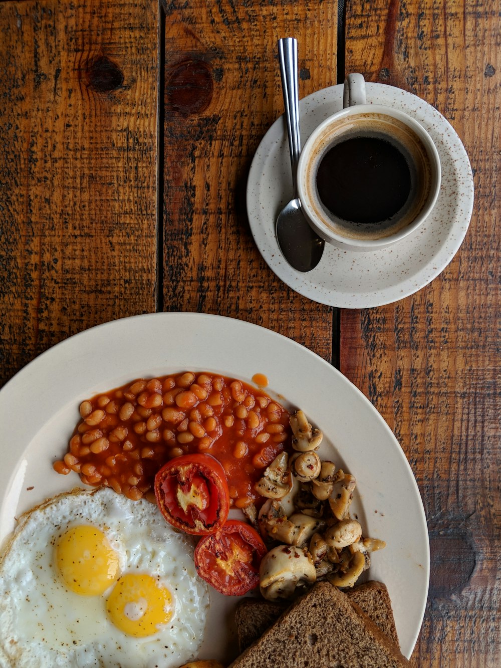 Schwarzer Kaffee in der Nähe von Sunny Side Up Ei mit geschnittenen Tomaten, Champignons und Brot auf dem Teller