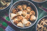 A collection of golden-brown samosas is arranged in a round metal tray placed on a green leafy surface. Other trays with various snacks surround the main tray. The handwritten label at the bottom likely indicates the type of food.