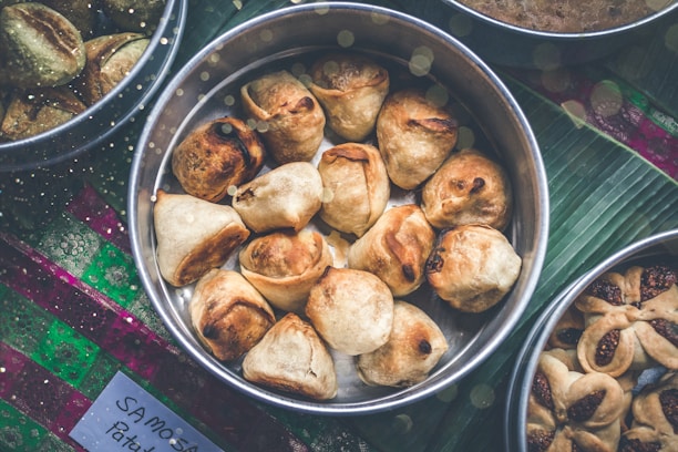 A collection of golden-brown samosas is arranged in a round metal tray placed on a green leafy surface. Other trays with various snacks surround the main tray. The handwritten label at the bottom likely indicates the type of food.
