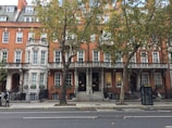 A row of elegant, traditional British townhouses with red brick facades and white window frames line the street. Trees with green foliage partially obscure the architecture, while a black bus stop is visible on the sidewalk.