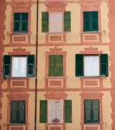 A building facade with a decorative painting that includes several windows, some of which are real with green shutters, and others painted alongside architectural embellishments. The background is a soft yellow with pinkish-orange accents.