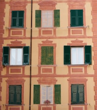 A building facade with a decorative painting that includes several windows, some of which are real with green shutters, and others painted alongside architectural embellishments. The background is a soft yellow with pinkish-orange accents.