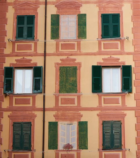 A building facade with a decorative painting that includes several windows, some of which are real with green shutters, and others painted alongside architectural embellishments. The background is a soft yellow with pinkish-orange accents.