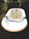 A vintage-style white bathtub with metal fixtures is centered in a rustic, possibly industrial setting. The bathtub has a wooden board placed across its width, which is likely used as a bath tray. The background includes weathered wooden doors, a barrel, and a sign with some text. The overall setting appears to be dimly lit, creating a nostalgic feel.