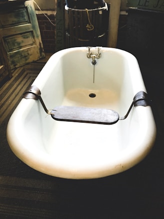 A vintage-style white bathtub with metal fixtures is centered in a rustic, possibly industrial setting. The bathtub has a wooden board placed across its width, which is likely used as a bath tray. The background includes weathered wooden doors, a barrel, and a sign with some text. The overall setting appears to be dimly lit, creating a nostalgic feel.