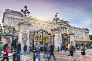 An iconic palace with grand architectural details featuring intricate gates adorned with gold and decorative crests. People walk by in the foreground, adding a sense of motion and activity. The sunlight highlights the building's facade.