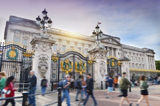 An iconic palace with grand architectural details featuring intricate gates adorned with gold and decorative crests. People walk by in the foreground, adding a sense of motion and activity. The sunlight highlights the building's facade.