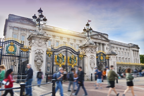 An iconic palace with grand architectural details featuring intricate gates adorned with gold and decorative crests. People walk by in the foreground, adding a sense of motion and activity. The sunlight highlights the building's facade.