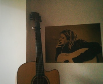 A guitar is resting against a wall next to a poster featuring a person smoking while holding a guitar. The poster has a sepia tone, and the signature on the guitar is visible.