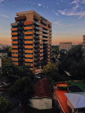 A multi-story residential building with a series of balconies is set against a backdrop of a blue sky with scattered clouds. The building features a combination of orange-brown and dark gray colors. Surrounding it are trees with lush green foliage, and in the foreground, there is a small structure with a sloped reddish-brown roof. The scene conveys a peaceful suburban setting with a hint of urban architecture.