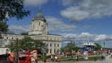 A historic stone building with a large clock tower and a domed roof stands prominently under a partly cloudy sky. People stroll through a vibrant park area with colorful flowers and a gazebo. Buses and tourists are visible in the foreground, suggesting a popular destination.