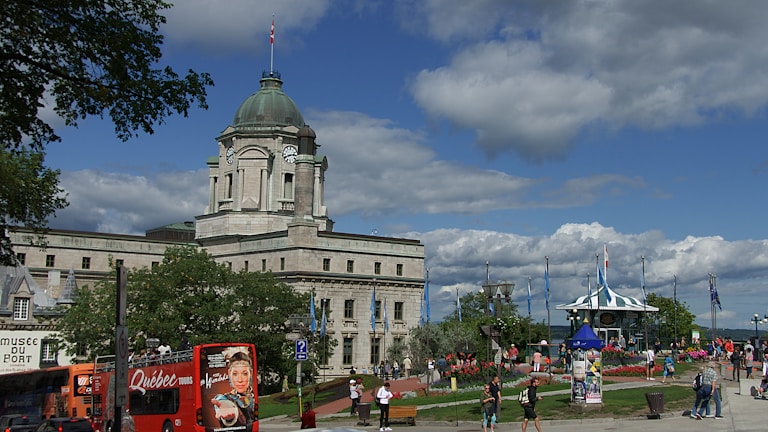 A historic stone building with a large clock tower and a domed roof stands prominently under a partly cloudy sky. People stroll through a vibrant park area with colorful flowers and a gazebo. Buses and tourists are visible in the foreground, suggesting a popular destination.