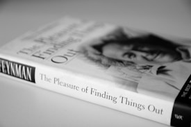A black and white photograph of a book titled 'The Pleasure of Finding Things Out' with a blurred portrait of a person on the cover. The book is lying on a flat surface, and the spine indicates the author as 'Feynman.'