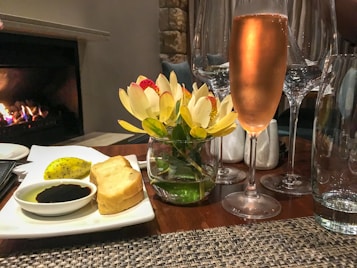 A cozy dining setting with a glass of ros&eacute; wine, surrounded by tall wine glasses on a wooden table. Beside it, a small vase holds vibrant flowers. A plate with bread slices, a greenish fruit, and a bowl of dark balsamic with olive oil sits nearby. In the background, a small fire burns, adding warmth to the atmosphere.