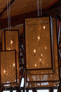 Hanging rectangular lanterns with intricate perforated designs give off a warm glow. The wooden frame and detailed patterns create a cozy and artistic atmosphere. The ceiling appears to be made of wood, adding to the rustic ambiance.
