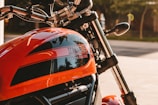 A close-up view of a motorcycle with an orange fuel tank featuring the brand name and decorative elements. The handlebars, mirrors, and parts of the chrome fork are visible, with a blurred background indicating an outdoor setting.