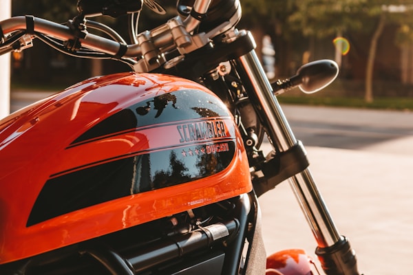 A close-up view of a motorcycle with an orange fuel tank featuring the brand name and decorative elements. The handlebars, mirrors, and parts of the chrome fork are visible, with a blurred background indicating an outdoor setting.