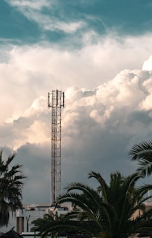 A tall communication tower stands prominently against a backdrop of expansive, fluffy clouds. The foreground features several palm trees and part of a building, contributing to a tropical or subtropical setting. The sky exhibits a range of blue to white hues, suggesting a calm weather condition.
