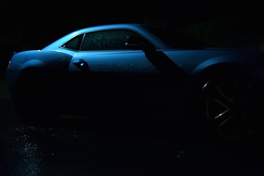A sleek, dark-colored car is illuminated with subtle blue lighting, highlighting its curves and details in a dimly lit environment. The surface of the car appears slightly wet, likely due to recent rain, which adds a reflective sheen.