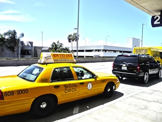 A yellow taxi cab is parked in front of a black SUV, both located near an airport terminal. The taxi displays a phone number and website for Yellow Cab Co. A Hertz rental truck is visible in the background, along with modern architecture and palm trees. The scene is well-lit with a clear blue sky.