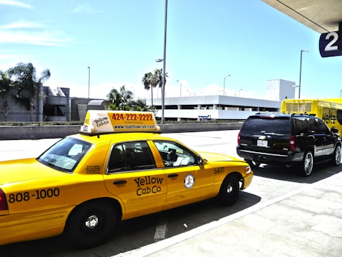 A yellow taxi cab is parked in front of a black SUV, both located near an airport terminal. The taxi displays a phone number and website for Yellow Cab Co. A Hertz rental truck is visible in the background, along with modern architecture and palm trees. The scene is well-lit with a clear blue sky.