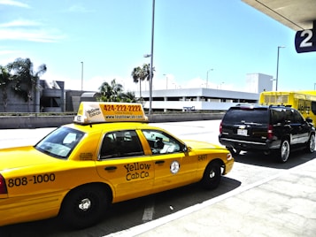 A yellow taxi cab is parked in front of a black SUV, both located near an airport terminal. The taxi displays a phone number and website for Yellow Cab Co. A Hertz rental truck is visible in the background, along with modern architecture and palm trees. The scene is well-lit with a clear blue sky.