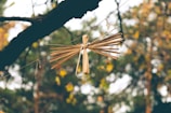 A handcrafted figure made from natural materials is suspended on a string, resembling an angel or bird. It's surrounded by a forest-like background with soft, blurred autumn foliage in yellows and greens.