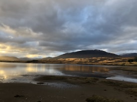 A tranquil landscape featuring a calm lake reflecting the golden hues of the setting or rising sun. Mountains in the background are partially shadowed by a dramatic sky filled with clouds. The foreground consists of a rocky and sandy shore.
