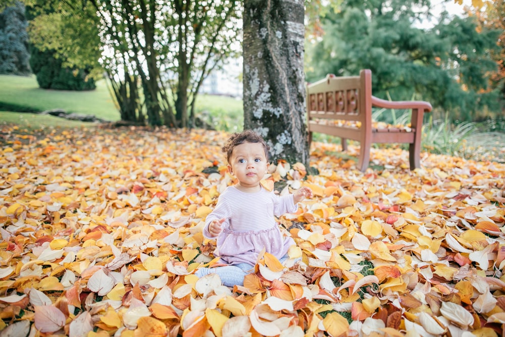 toddler sitting on leaf-covered ground near bench during daytime