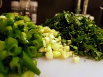 Green onions, garlic, and parsley are chopped finely and arranged on a white cutting board in a well-lit kitchen setting. In the background, blurred spice jars are visible.
