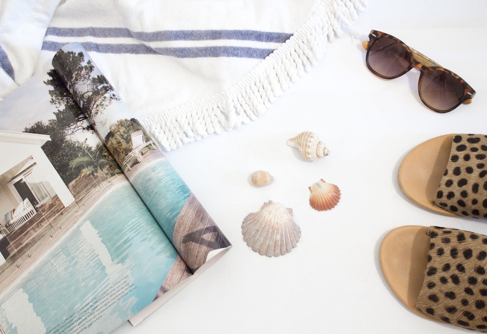 opened magazine beside seashells, slide sandals, sunglasses and towel on white surface