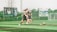A group of young men are playing soccer on a sports field with artificial turf. The players are in motion, chasing after the ball, displaying a sense of dynamic action and teamwork. In the background, there is a fence with several large advertisement banners featuring logos and text.