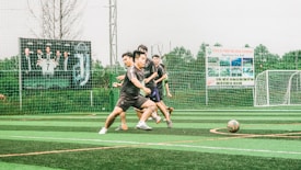 A group of young men are playing soccer on a sports field with artificial turf. The players are in motion, chasing after the ball, displaying a sense of dynamic action and teamwork. In the background, there is a fence with several large advertisement banners featuring logos and text.