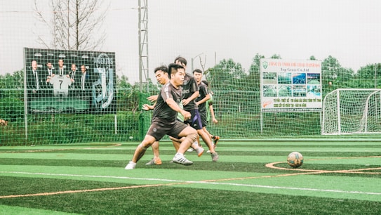 A group of young men are playing soccer on a sports field with artificial turf. The players are in motion, chasing after the ball, displaying a sense of dynamic action and teamwork. In the background, there is a fence with several large advertisement banners featuring logos and text.
