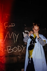 A person stands in front of a dark wall with the words 'Find My Body' written in chalk. They wear a blue patterned hospital gown over casual clothing, covering their face with one hand. A small stuffed toy is visible near their arm. The image is lit with a red glow from the left, adding a dramatic effect.