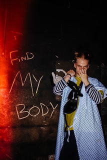 A person stands in front of a dark wall with the words 'Find My Body' written in chalk. They wear a blue patterned hospital gown over casual clothing, covering their face with one hand. A small stuffed toy is visible near their arm. The image is lit with a red glow from the left, adding a dramatic effect.