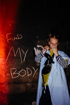 A person stands in front of a dark wall with the words 'Find My Body' written in chalk. They wear a blue patterned hospital gown over casual clothing, covering their face with one hand. A small stuffed toy is visible near their arm. The image is lit with a red glow from the left, adding a dramatic effect.