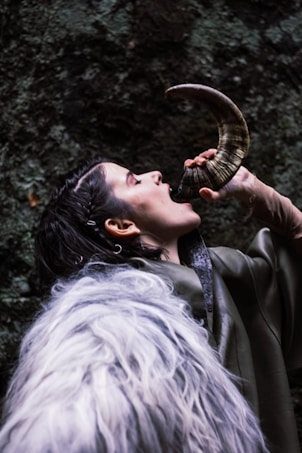 A person with dark hair braided on the side is blowing into a curved horn. They appear to be wearing a grayish-green cloak, and a white fur garment is visible on their back. The background is dark and textured, resembling a forest or rocky landscape.