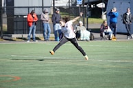 A person is mid-air on a sports field, appearing to be involved in an athletic activity. Several spectators are on the sidelines, some seated and others standing, with one individual capturing the event with a camera. The background includes a fence and various casual observers, some of whom are wearing winter clothing.