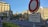 A city scene featuring a street sign with French text in the foreground, set against a backdrop of old-style buildings under a clear blue sky. There's a distant clock tower, a few pedestrians, and minimal traffic. The intersection is surrounded by various traffic signs and landscaped greenery.