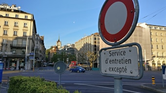 A city scene featuring a street sign with French text in the foreground, set against a backdrop of old-style buildings under a clear blue sky. There's a distant clock tower, a few pedestrians, and minimal traffic. The intersection is surrounded by various traffic signs and landscaped greenery.