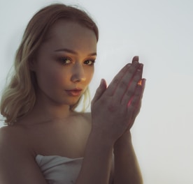 A young woman with blonde hair stands with her hands clasped, creating a contemplative and introspective pose. Soft lighting highlights her facial features, and the neutral background emphasizes a serene atmosphere.