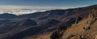 A vast volcanic landscape with rugged terrain, featuring rocky slopes, dark volcanic cones, and expansive valleys. The horizon is adorned with fluffy white clouds floating above the dark, craggy mountains. The play of light and shadow across the landscape highlights the dramatic contours and textures of the volcanic region.