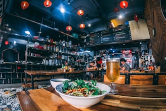 A cozy restaurant interior with wooden tables and vibrant red lanterns hanging from the ceiling. A bowl of food garnished with greens and a chilled glass of beer are placed on the table in the foreground. Shelves stocked with various jars and utensils line the walls, creating a busy yet inviting atmosphere. In the background, patrons are seated at tables, engaged in quiet conversation under the warm lighting.