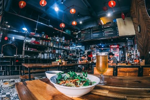 A cozy restaurant interior with wooden tables and vibrant red lanterns hanging from the ceiling. A bowl of food garnished with greens and a chilled glass of beer are placed on the table in the foreground. Shelves stocked with various jars and utensils line the walls, creating a busy yet inviting atmosphere. In the background, patrons are seated at tables, engaged in quiet conversation under the warm lighting.