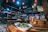 A cozy restaurant interior with wooden tables and vibrant red lanterns hanging from the ceiling. A bowl of food garnished with greens and a chilled glass of beer are placed on the table in the foreground. Shelves stocked with various jars and utensils line the walls, creating a busy yet inviting atmosphere. In the background, patrons are seated at tables, engaged in quiet conversation under the warm lighting.
