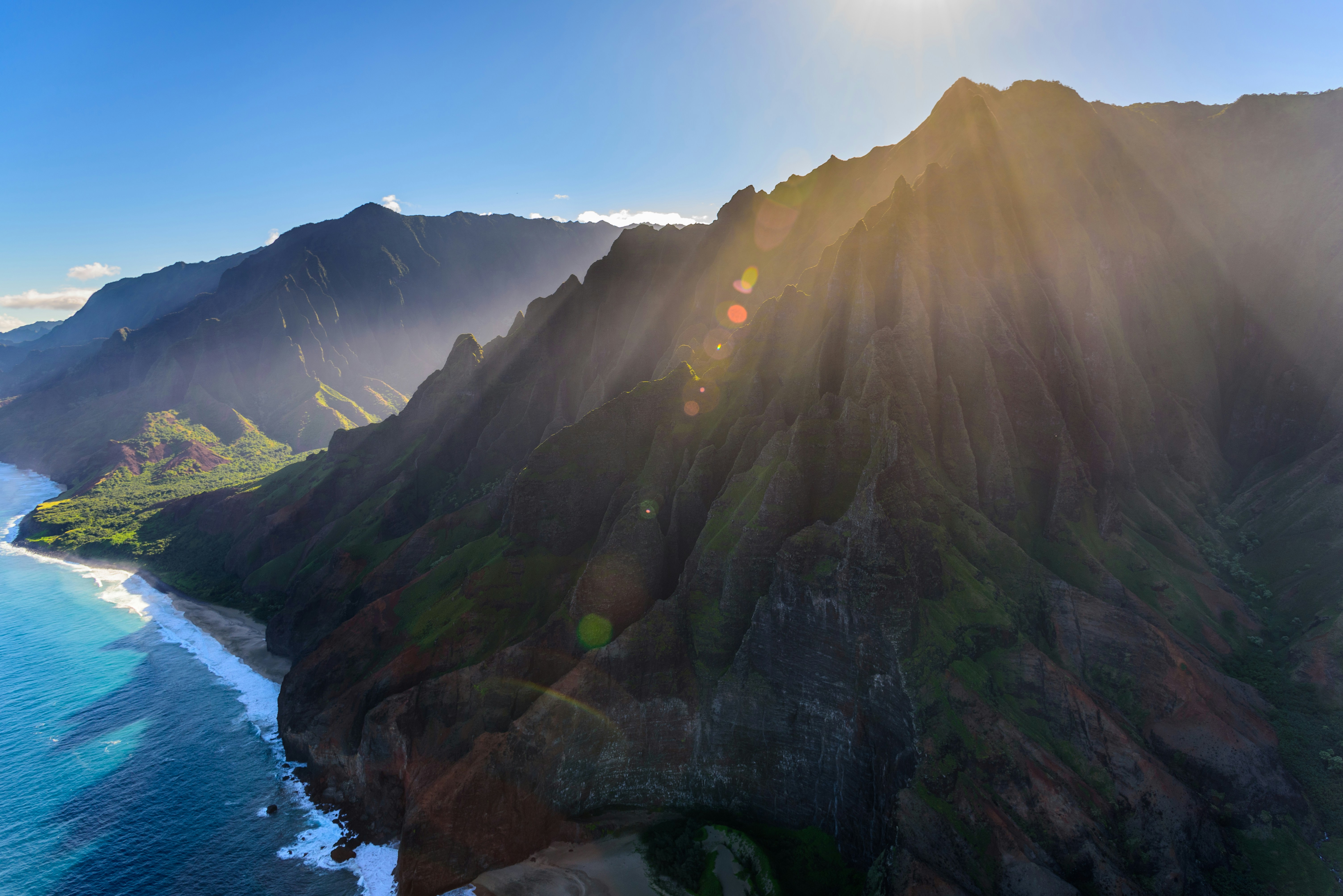 aerial view of mountain beside ocean, We were flying over Kauai on a helicopter with doors off and as we got to the Na’Pali coast, the iconic cathedrals were bathed in sunlight. I was so fascinated by this and wanted to capture it before the helicopter moved out of the scene. It was a very heavenly sight and I did not want to miss it. My camera went click.