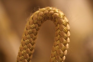 A close-up of a woven rope featuring a braided pattern with a looped curve in the foreground. The rope appears to be made of synthetic material with a golden tan color. The background is softly blurred, contributing to the focus on the rope's texture and intricate weave.
