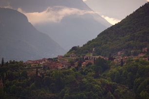 A lush green valley enveloped by tall mountains is dotted with quaint houses. The village is nestled amidst dense foliage, with a few towers and buildings standing out. Mist hovers over the mountaintops, adding a serene and tranquil atmosphere to the landscape.
