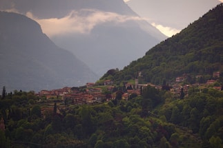 A lush green valley enveloped by tall mountains is dotted with quaint houses. The village is nestled amidst dense foliage, with a few towers and buildings standing out. Mist hovers over the mountaintops, adding a serene and tranquil atmosphere to the landscape.